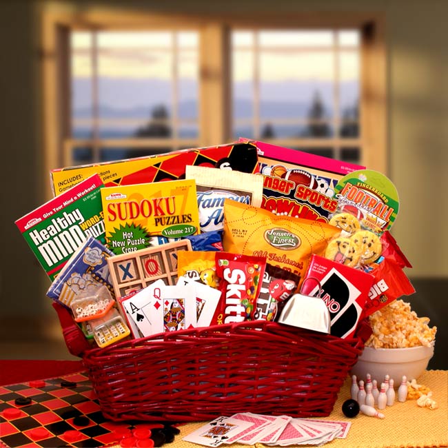 Large red wicker basket is filled with a Checkers game set, Word search puzzle, book, Sudoku puzzle book, Crossword puzzle book, Tic-tac-toe wooden game set, Word Boggle game cube, Finger bowling game, Mini travel game, Cowbell noise maker, UNO card game,Casino-style playing cards and a variety of snack and sweets
